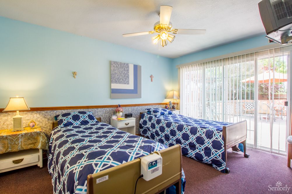 A well-lit nursing home bedroom with twin beds, patterned bedding, and a ceiling fan.