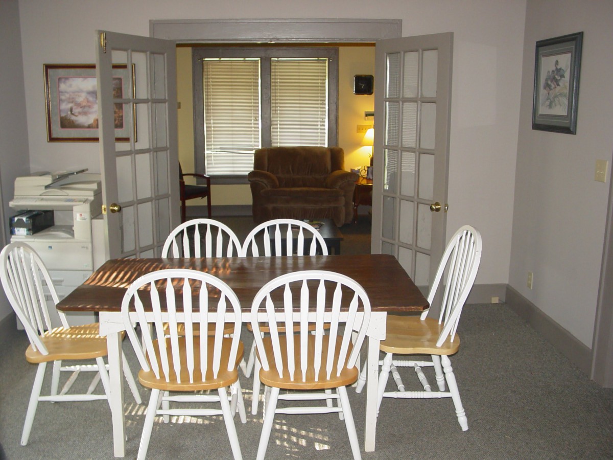 A dining area with a wooden table and white chairs, leading to a living room through French doors.