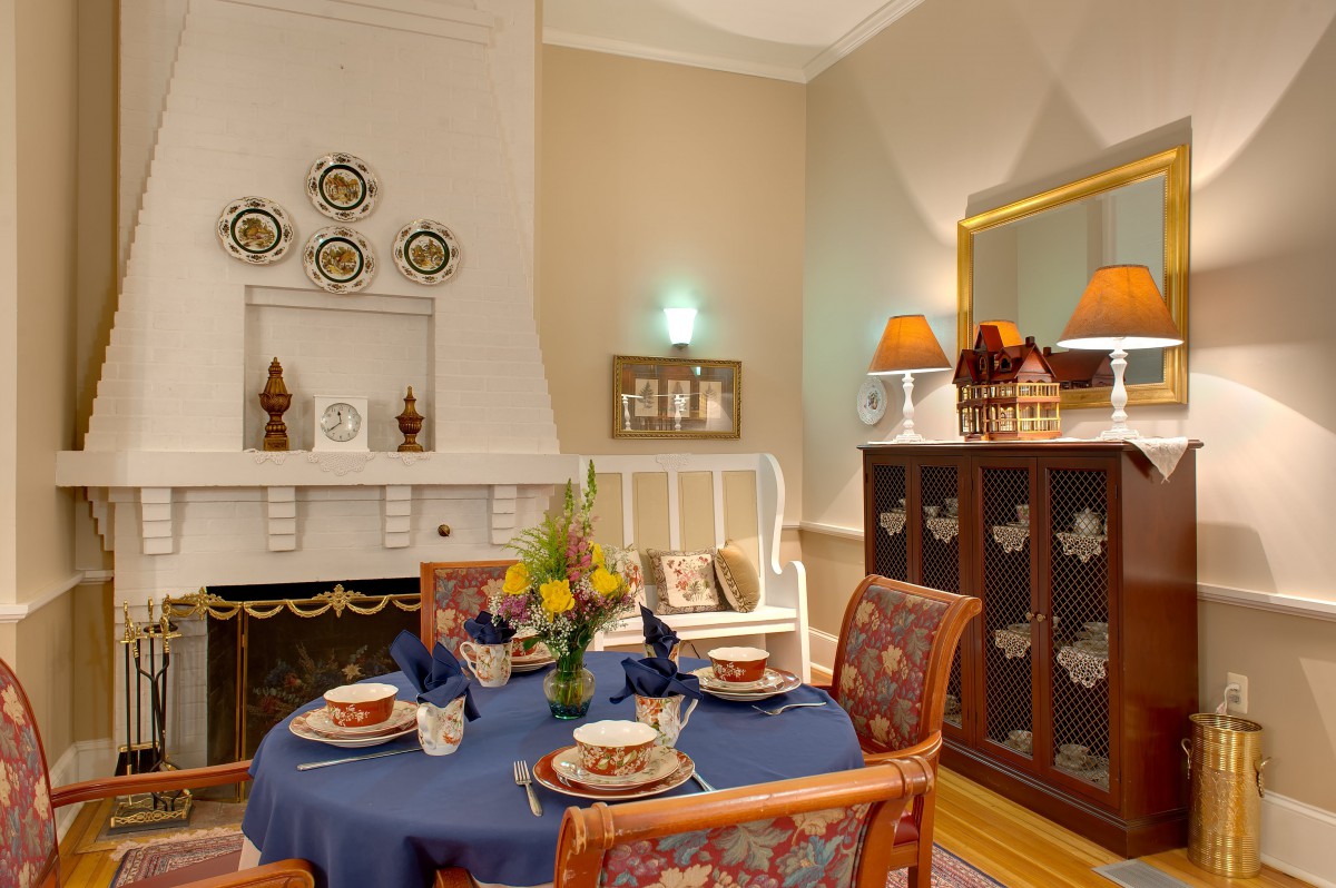 A warm and inviting dining room featuring a round table with a blue tablecloth, surrounded by patterned chairs. The decor includes elegant wall art, a decorative cabinet with lamps, and a cozy fireplace.