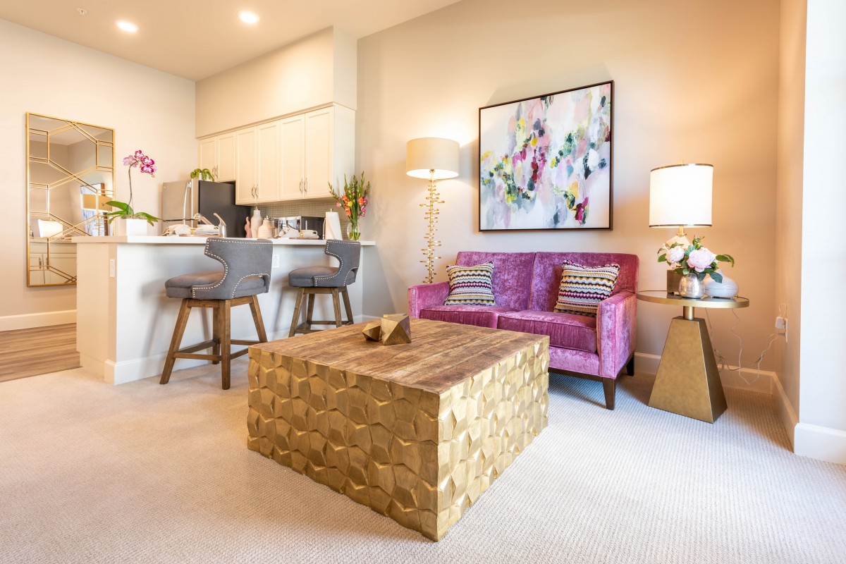 A modern living room with a pink sofa, abstract wall art, and a wooden coffee table, adjacent to an open kitchen area with bar stools.
