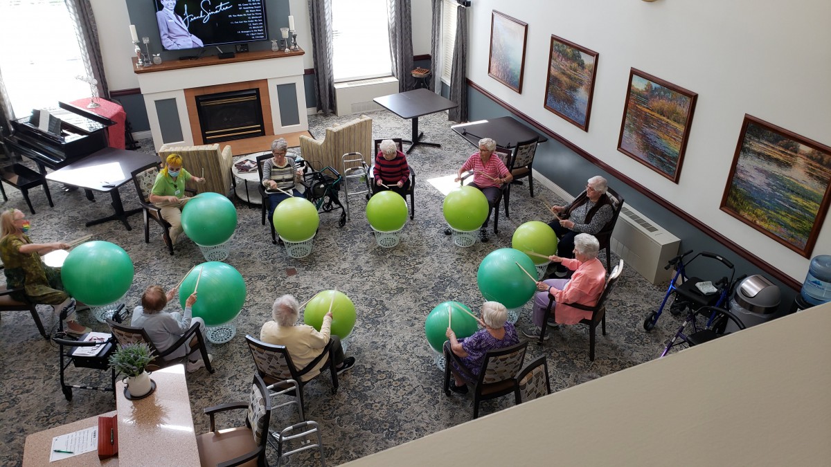 A group of seniors participates in a seated exercise class with green fitness balls, arranged in a circle.