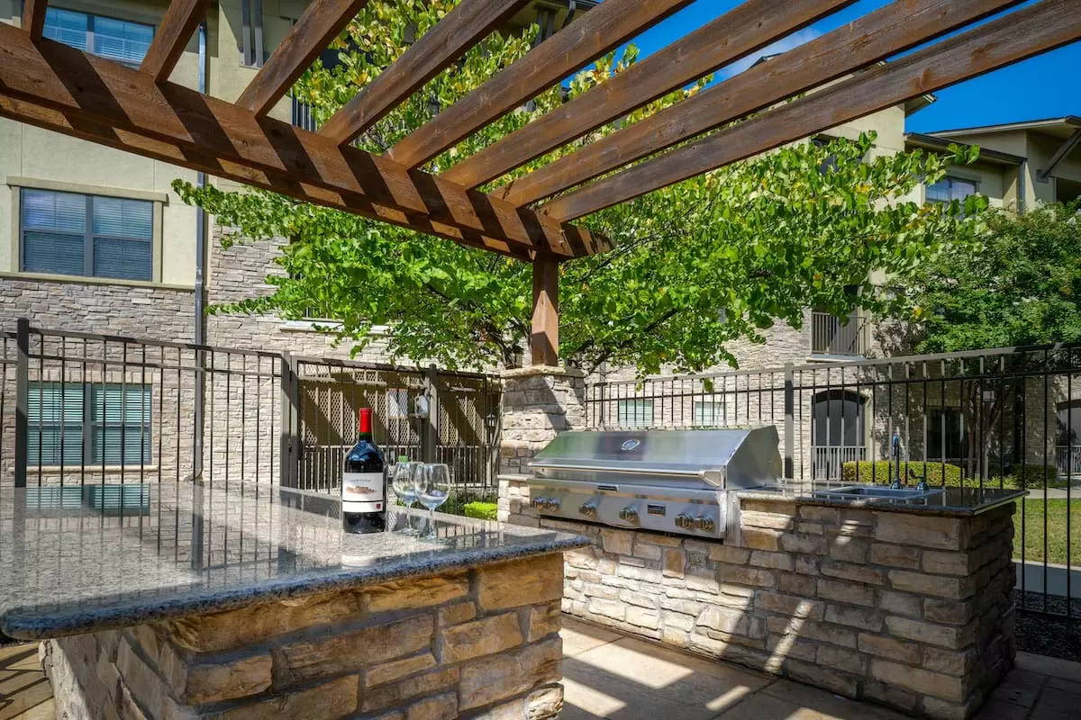 A sunny outdoor kitchen with a grill under a wooden pergola, surrounded by a stone counter and shaded by trees.
