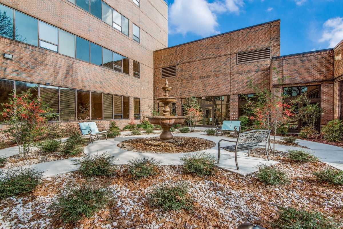 A peaceful courtyard with a central fountain, surrounded by benches and chairs, adjacent to a modern building.