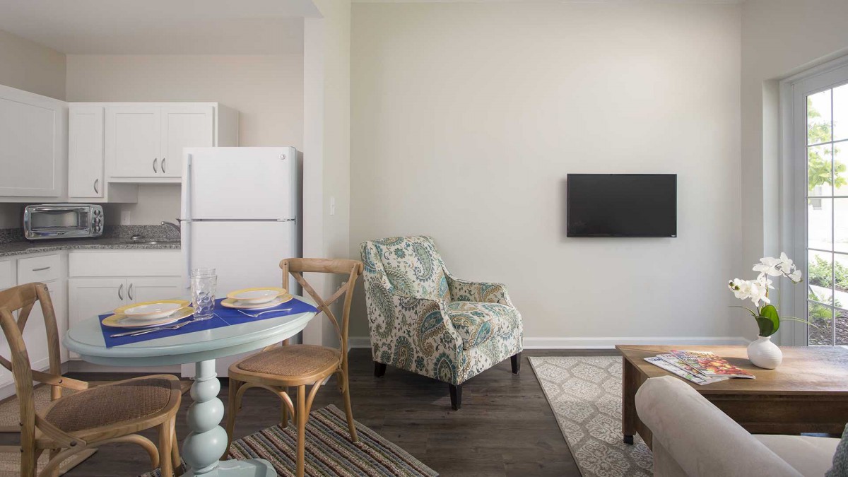 A cozy living area with a kitchen, featuring an armchair, round dining table, and wall-mounted television.