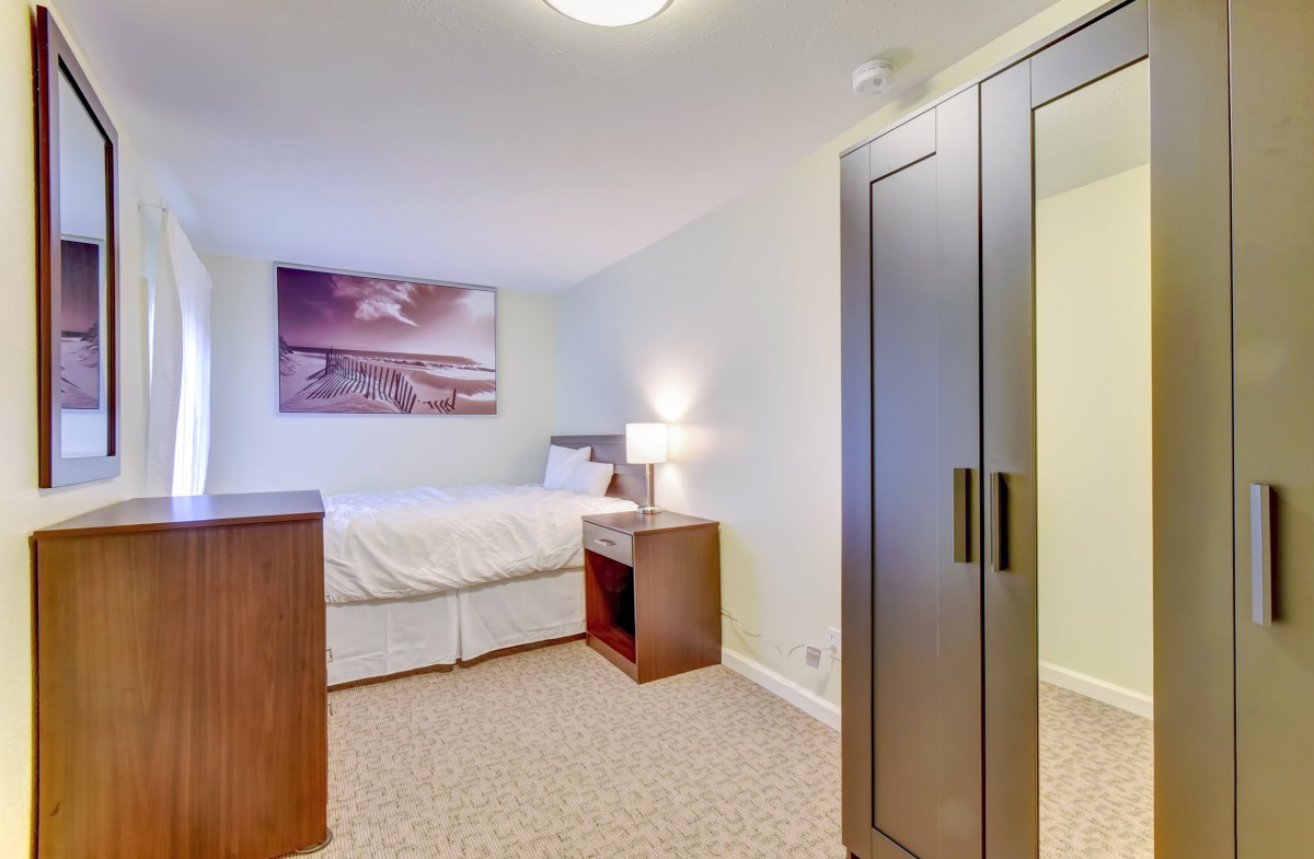 A cozy bedroom featuring a bed, wardrobe, and bedside lamp, with artwork on the wall.