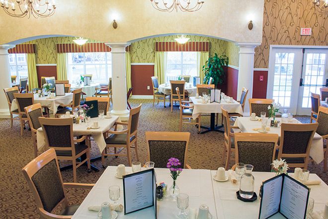 A well-decorated dining room with neatly arranged tables and chairs, featuring soft lighting and large windows.