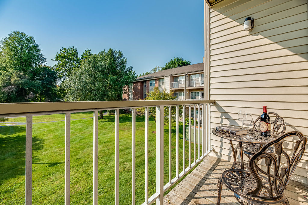 A sunny balcony with a small table and chairs overlooking a green lawn in an apartment complex.
