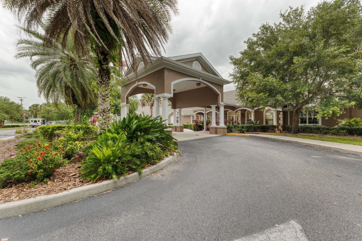 A building entrance with a circular driveway surrounded by lush landscaping and palm trees under a cloudy sky.