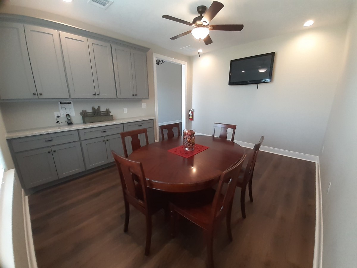A dining area with a wooden table and chairs, equipped with a ceiling fan and wall-mounted television.