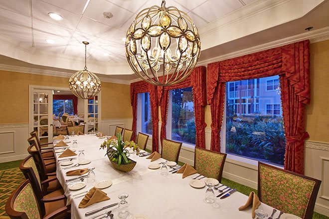 An elegantly decorated dining room with a long table set for a formal gathering, featuring large chandeliers and red curtains framing the windows.