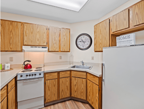 A compact kitchen with wood cabinets, featuring an oven, refrigerator, sink, and a wall clock.