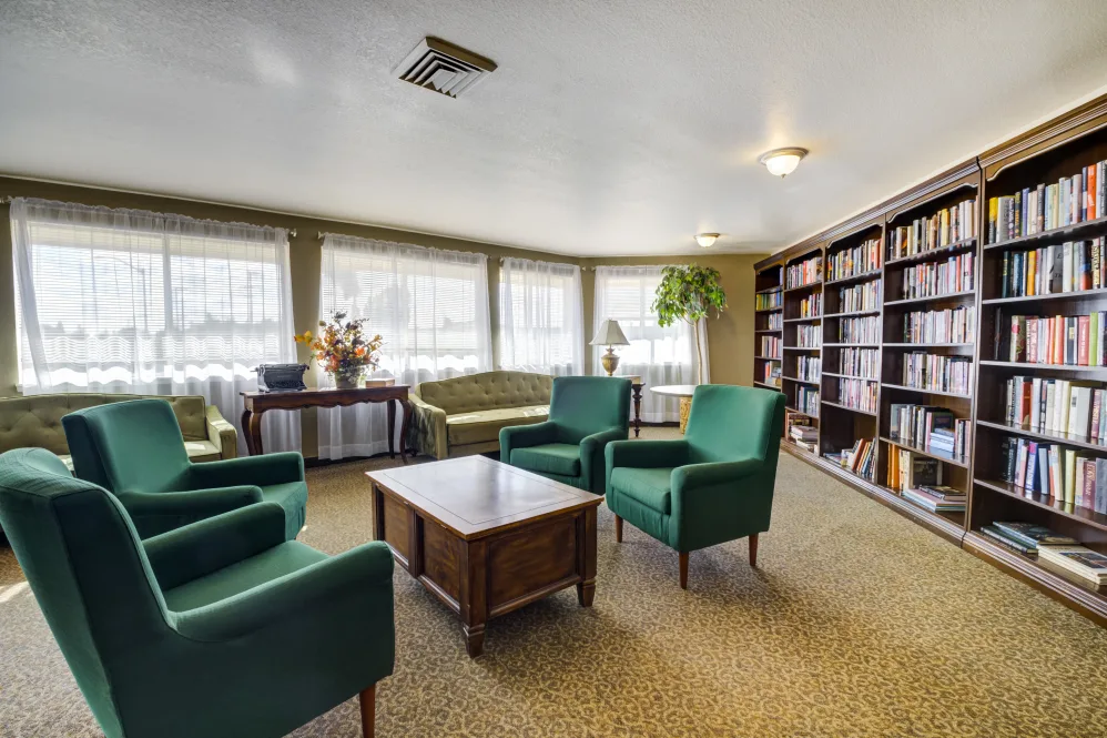 A cozy living room with armchairs and bookshelves, featuring a coffee table and large windows providing ample natural light.