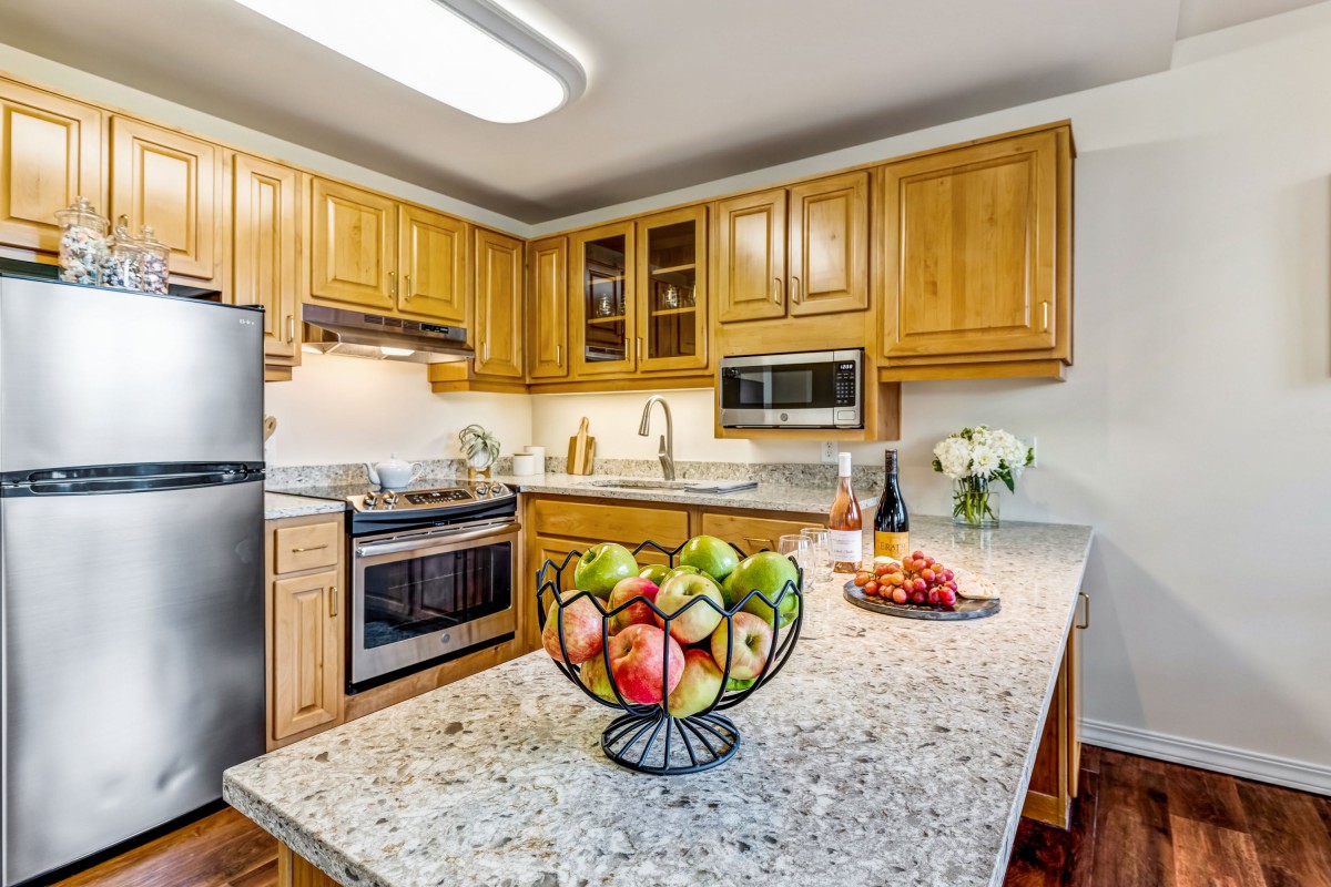A modern kitchen with wooden cabinets, granite countertops, and a fruit basket on the island.