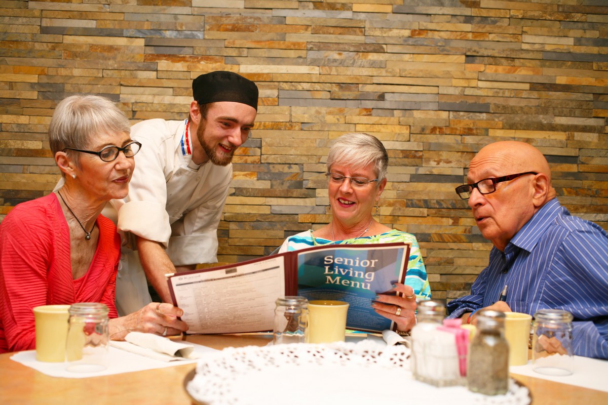 A group of elderly people reviewing a menu with a waiter in a restaurant.