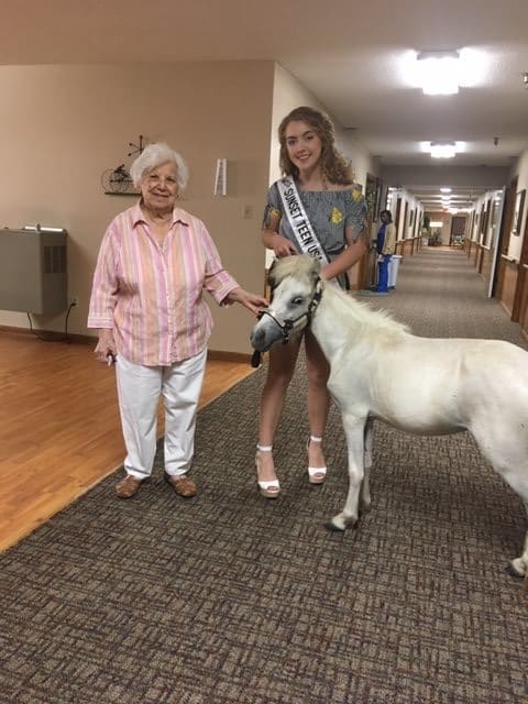 A young woman with a sash holds the lead of a miniature horse next to an elderly woman in a hallway.
