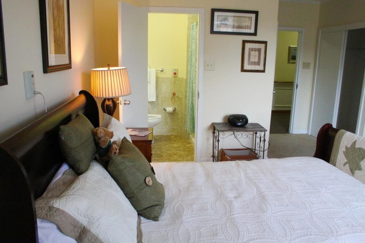 A cozy bedroom with an adjoining bathroom, featuring a neatly made bed adorned with pillows and a bedside lamp.