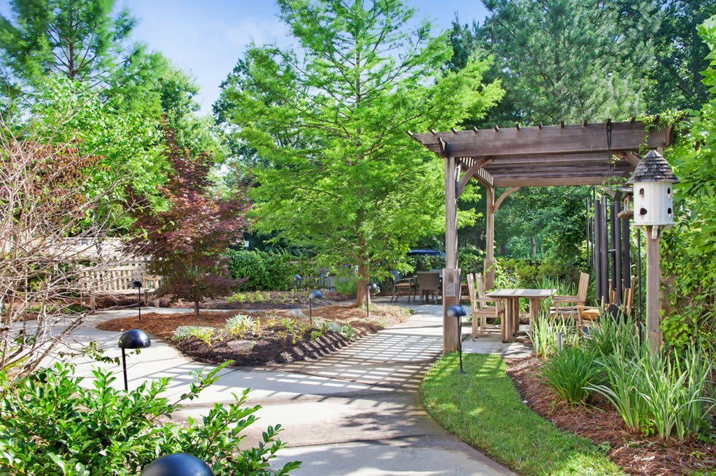 A beautiful garden with a winding pathway, surrounded by lush greenery and trees, featuring a pergola with outdoor seating.