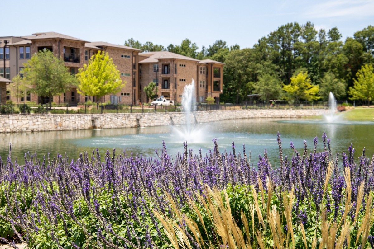 A picturesque view of an apartment complex with a fountain in a pond, surrounded by lush greenery and flowers.