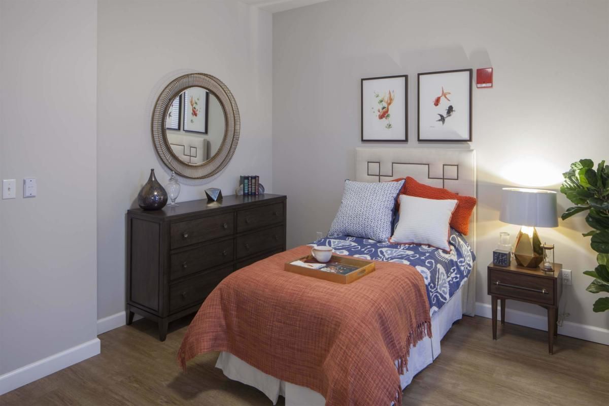 A cozy bedroom with a single bed adorned with colorful pillows and a throw blanket, featuring wall art, a mirror, and a dark wooden dresser.