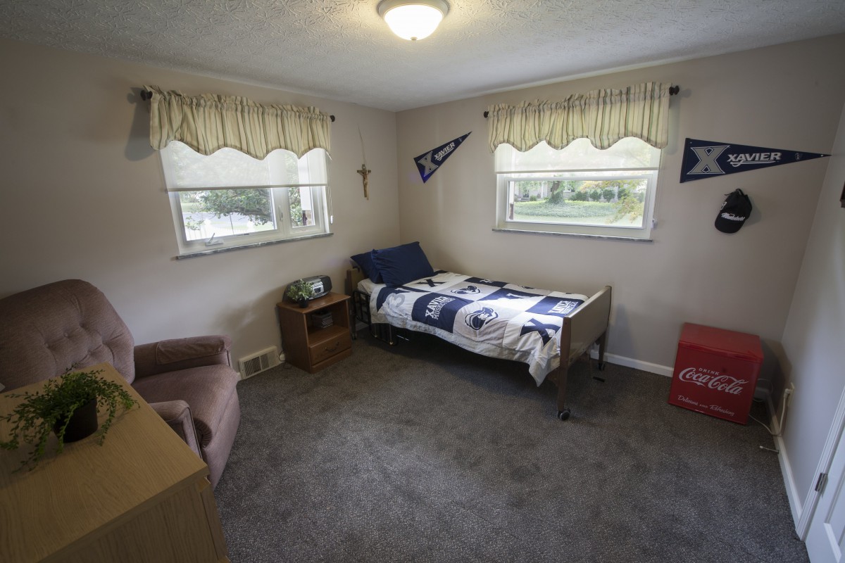 A small bedroom with a single bed, carpeted floor, and two windows adorned with curtains. The room features decorative pennants and wall art.