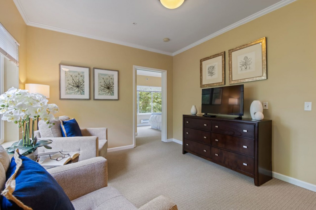A cozy living room with a beige color scheme, featuring a sofa, wall art, a television on a dark wooden dresser, and carpeted flooring.