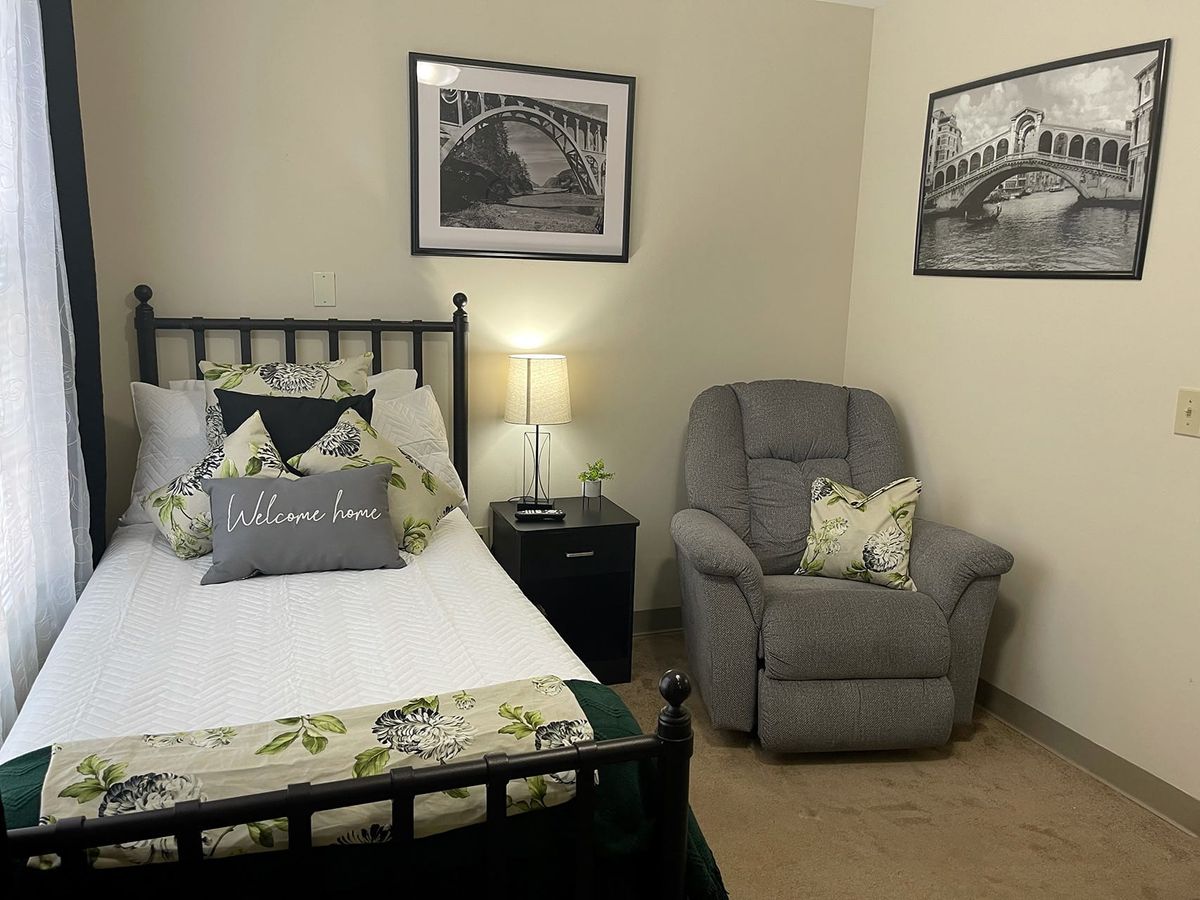 A cozy bedroom with a bed, nightstand, and armchair, featuring wall art and decorative pillows.