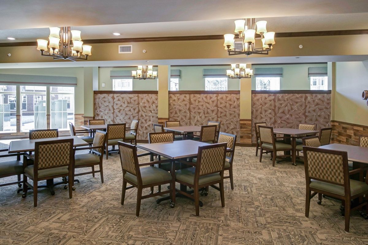A spacious dining area with multiple tables and chairs, decorated with elegant chandeliers and carpeted flooring.