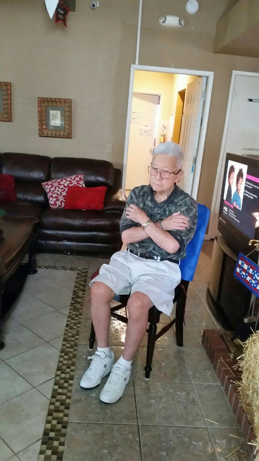 An elderly person sitting in a living room with crossed arms, watching television.