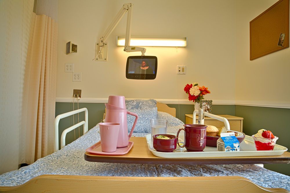 A hospital room with a bed, a meal tray with cups and flowers, and a wall-mounted television.