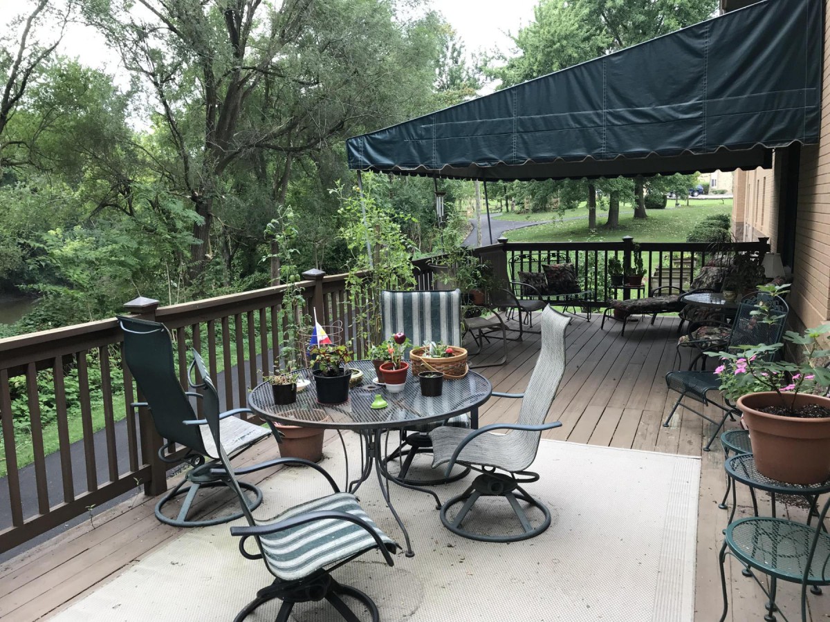 A cozy outdoor deck area with a table, chairs, and an awning surrounded by lush greenery.