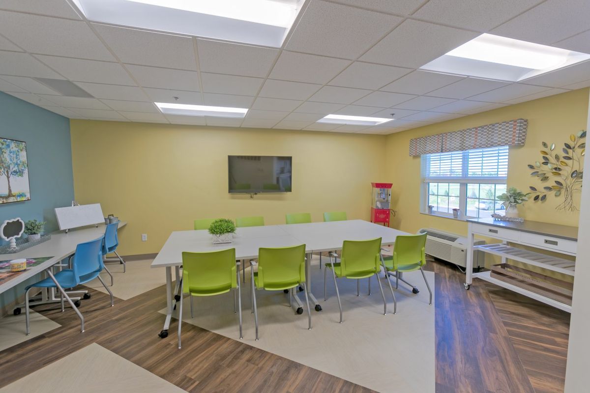 A bright and modern meeting room with green chairs around a large conference table, featuring a wall-mounted television and ample natural light through the windows.
