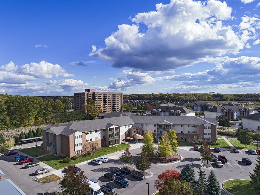 Aerial view of an apartment complex with a parking lot, surrounded by trees and under a cloudy blue sky.