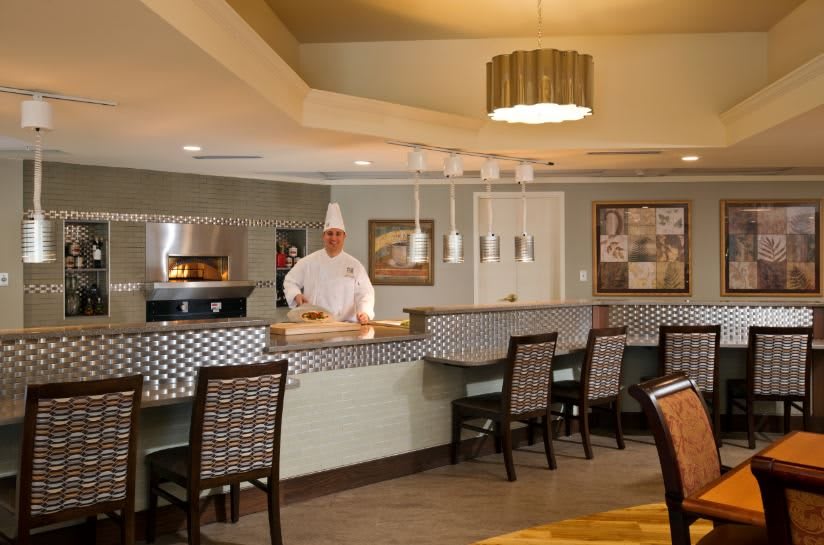 A chef stands behind the counter in a restaurant kitchen with dining area seating.