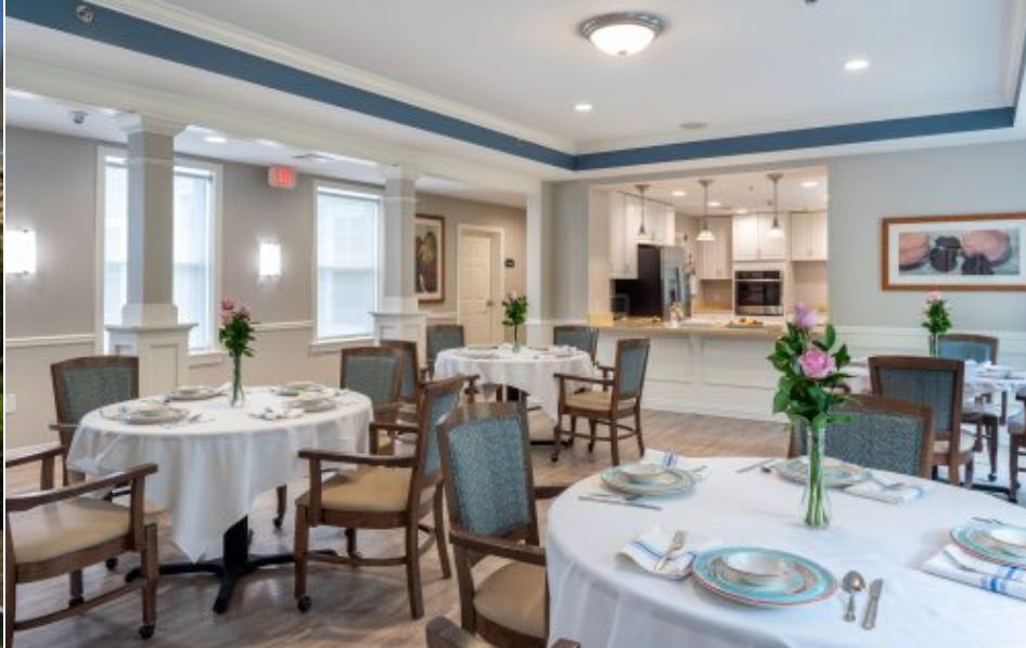An elegantly set dining area with neatly arranged tables and chairs, featuring floral centerpieces and a view of the kitchen.