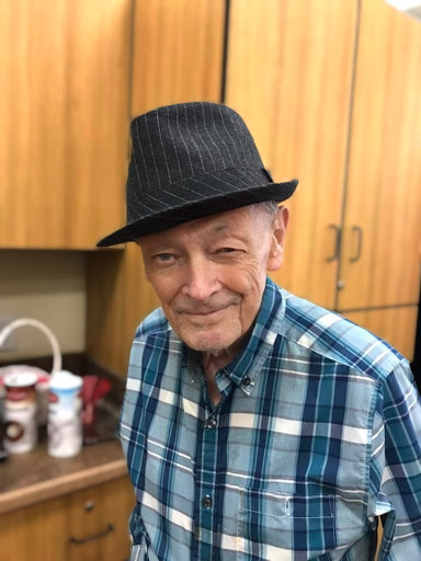 An elderly man wearing a stylish black hat and a plaid shirt, smiling warmly.