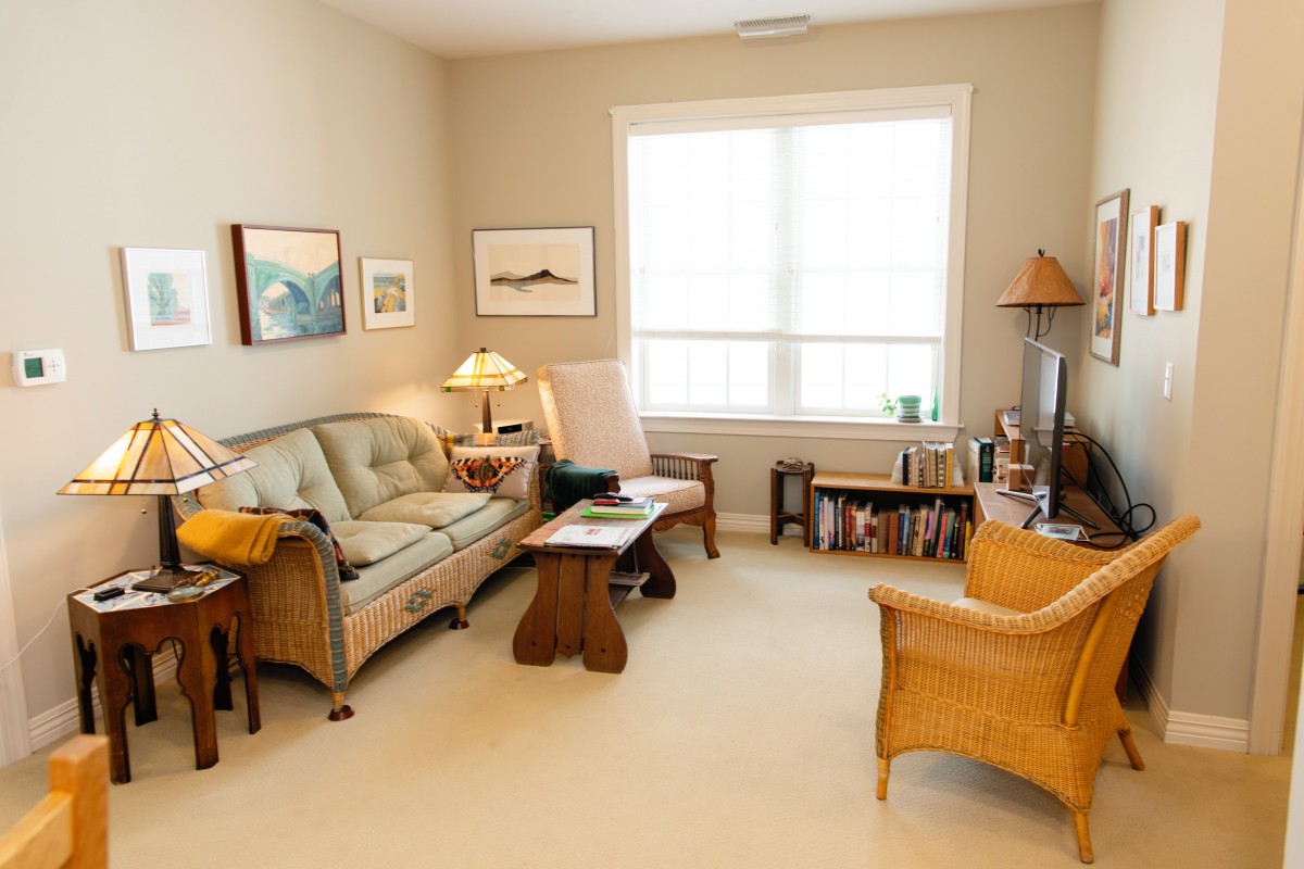 A cozy living room with a sofa, armchair, and coffee table, decorated with artwork and a lamp.