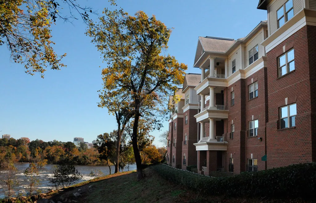 A view of an apartment building with balconies overlooking a river, surrounded by trees with autumn foliage.