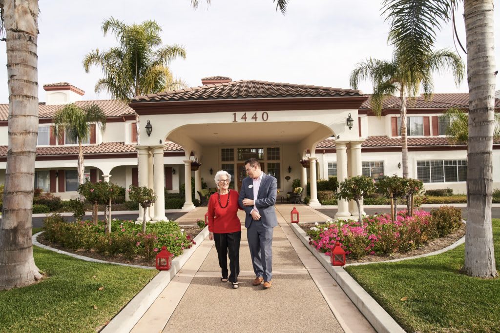 A senior woman and a man walking together on a pathway in front of a residential building with well-maintained landscaping.
