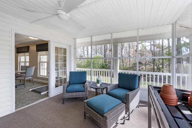 A cozy sunroom featuring blue-cushioned patio furniture, including two armchairs and a small table, with a ceiling fan overhead and large windows providing natural light.