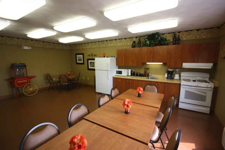 A communal kitchen and dining area with tables, chairs, a refrigerator, microwave, and decorative flowers.
