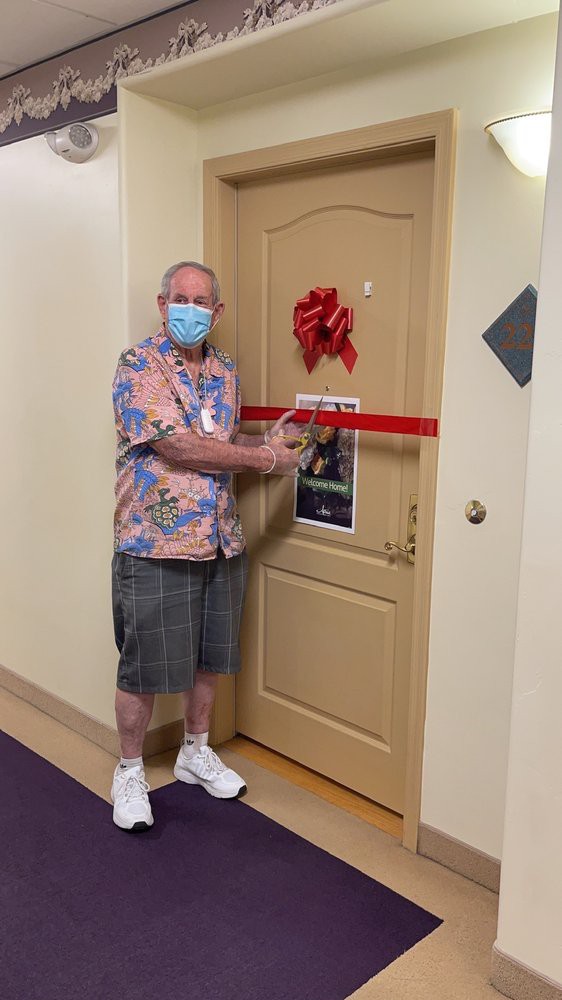 A man wearing a mask is cutting a red ribbon in front of a door decorated with a bow.