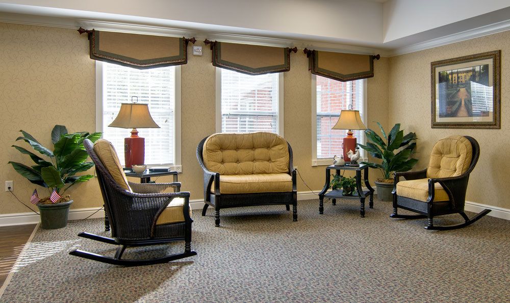 A cozy living room with a sofa, chairs, lamps, and indoor plants, set against a backdrop of large windows.
