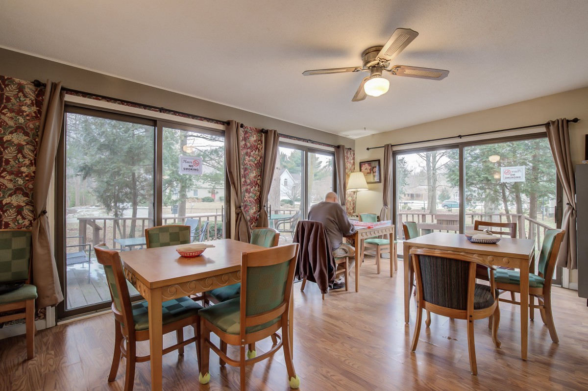 A cozy dining area with multiple tables and chairs, featuring large windows and a ceiling fan.