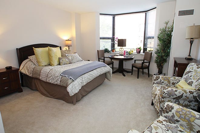 A cozy bedroom with a comfortable bed, natural lighting from large windows, and elegant furnishings.