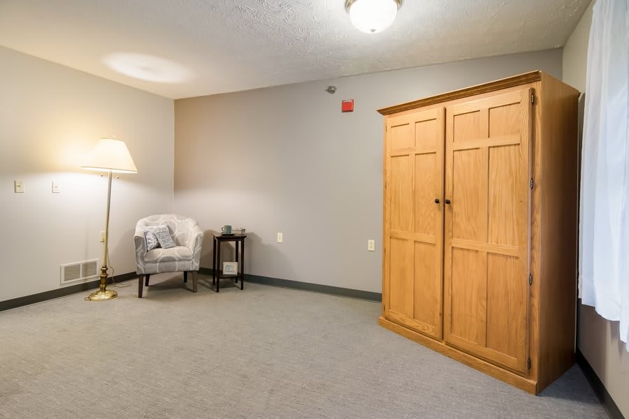 A minimalist room featuring a wooden wardrobe, an armchair beside a floor lamp, and neutral-colored walls and carpet.