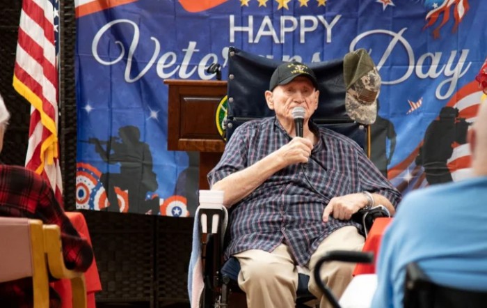 An elderly veteran speaks at a Veterans Day celebration, holding a microphone in front of an American flag backdrop.