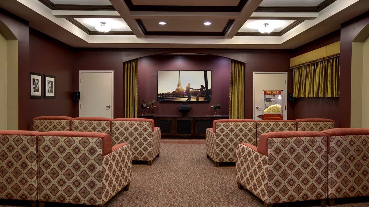 A cozy theater room with multiple patterned chairs facing a large screen, surrounded by warm decorative walls and lighting.