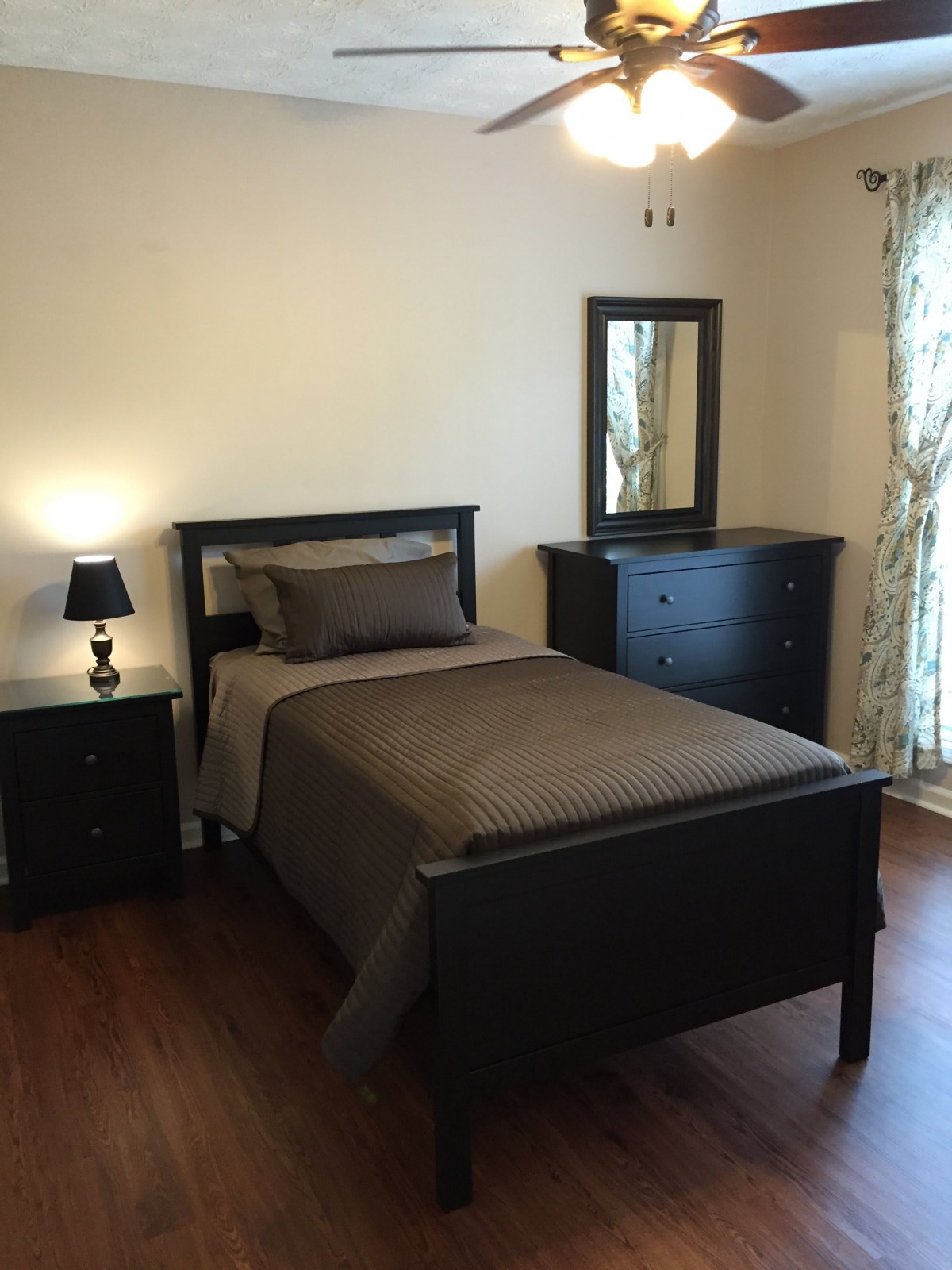 A minimalist bedroom with a single bed, dresser, and nightstand in dark wood tones and neutral bedding.