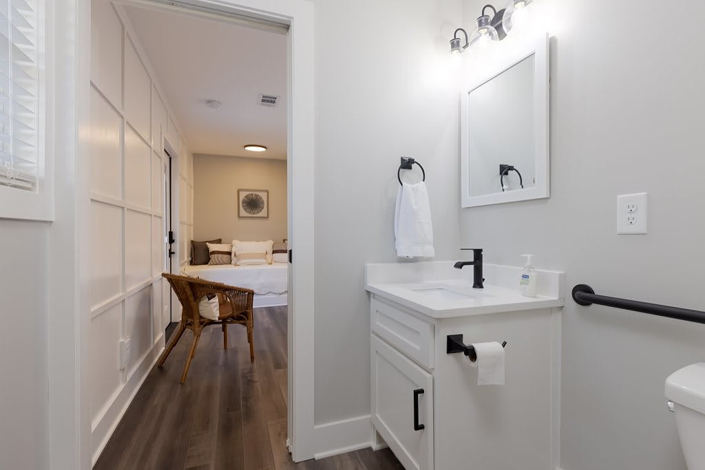 A modern, minimalist bathroom with a white vanity and wood flooring opens into a cozy room.