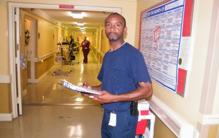 A healthcare worker in scrubs holds a clipboard in a hospital hallway.